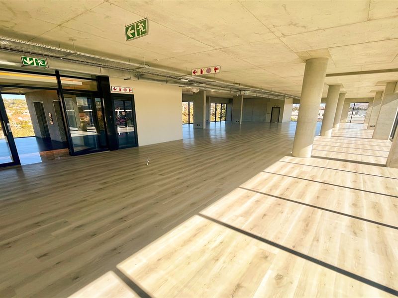 A spacious, modern open-plan office area featuring exposed concrete ceilings, large windows with natural light, and high-quality laminate flooring.