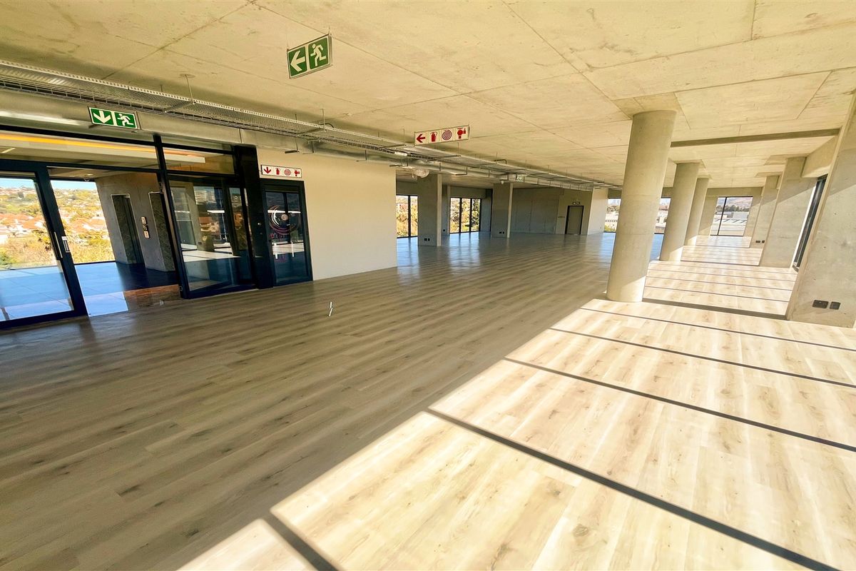 A spacious, modern open-plan office area featuring exposed concrete ceilings, large windows with natural light, and high-quality laminate flooring.