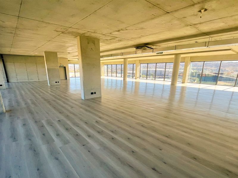 A spacious, open-plan office area featuring modern laminate flooring, exposed concrete ceilings, and expansive floor-to-ceiling windows providing ample natural light.