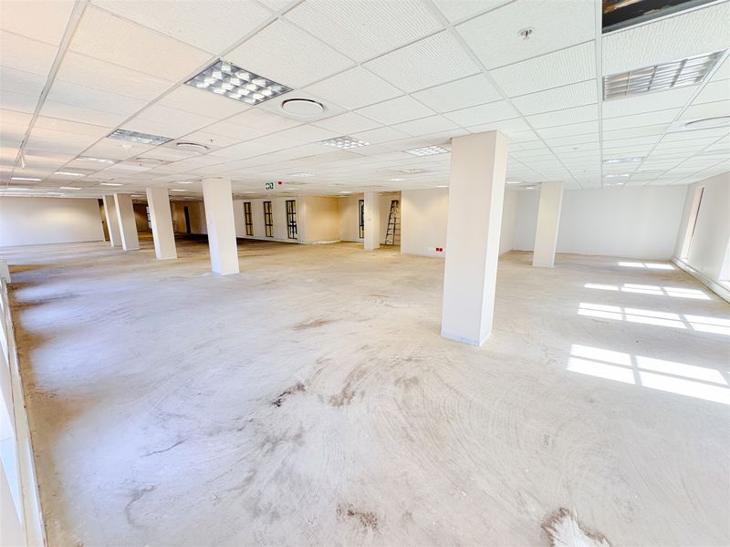 A large, open-plan office space featuring a suspended ceiling, recessed lighting, and structural columns, currently awaiting final flooring installation.