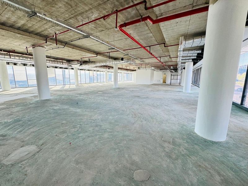 A large, open-plan office space in shell condition featuring exposed concrete ceilings, industrial piping, and expansive floor-to-ceiling windows providing ample natural light.