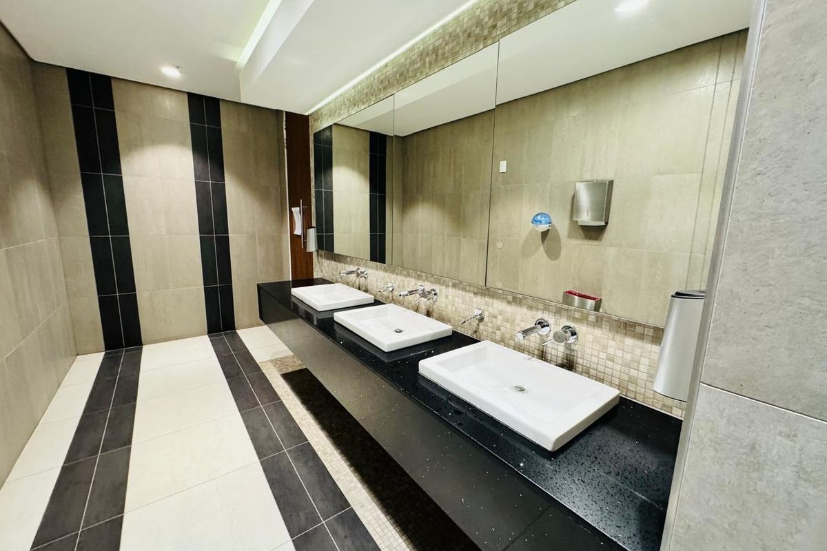 A modern, well-maintained office bathroom featuring a triple vanity with wall-mounted faucets, large mirror, and contemporary tiled finishes.