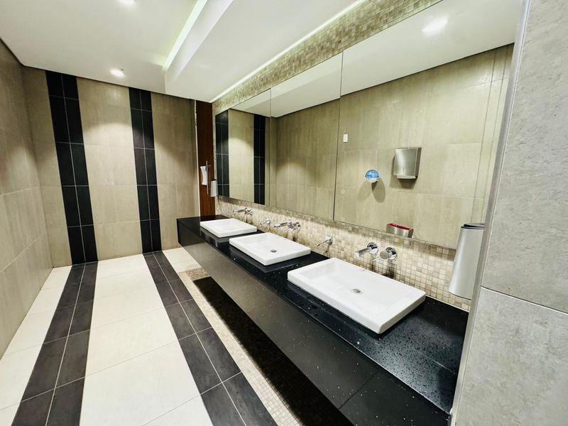 A modern, well-maintained office bathroom featuring a triple vanity with wall-mounted faucets, large mirror, and contemporary tiled finishes.