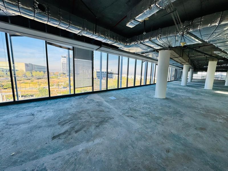 An expansive, open-plan office space in shell condition, featuring floor-to-ceiling windows, exposed industrial ceiling services, and structural columns with views of the surrounding area.