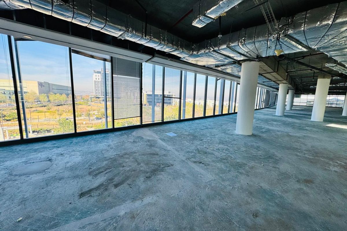 An expansive, open-plan office space in shell condition, featuring floor-to-ceiling windows, exposed industrial ceiling services, and structural columns with views of the surrounding area.