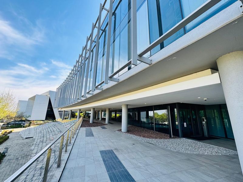 Modern office building exterior featuring a glass curtain wall, metal facade detailing, and a spacious paved entrance walkway with structural columns.