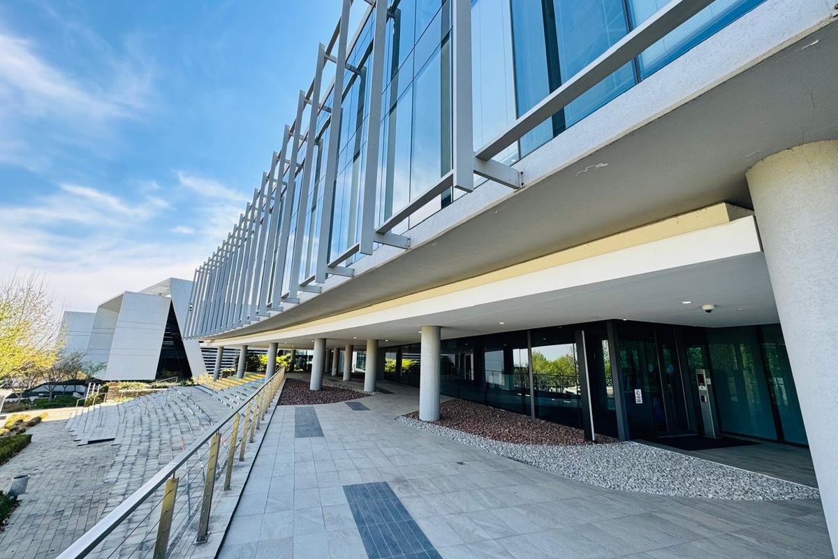 Modern office building exterior featuring a glass curtain wall, metal facade detailing, and a spacious paved entrance walkway with structural columns.