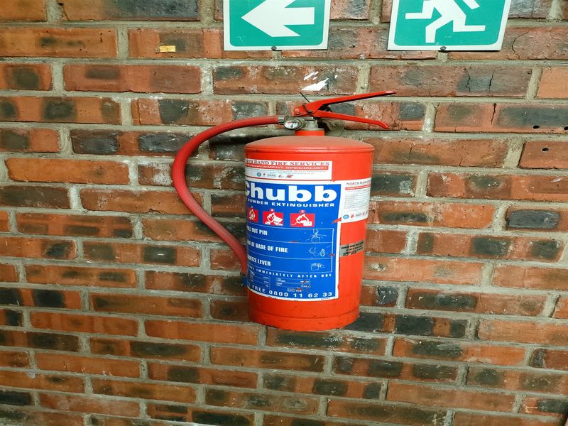 A wall-mounted fire extinguisher and emergency exit signage installed on an exposed brick wall within an industrial utility space.