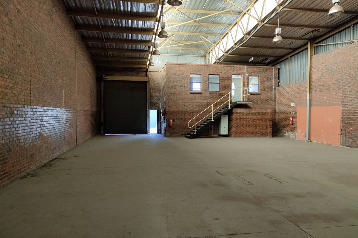 Spacious industrial warehouse featuring exposed brick walls, high-clearance steel truss roofing, concrete flooring, and a roller shutter door with an integrated mezzanine office area.