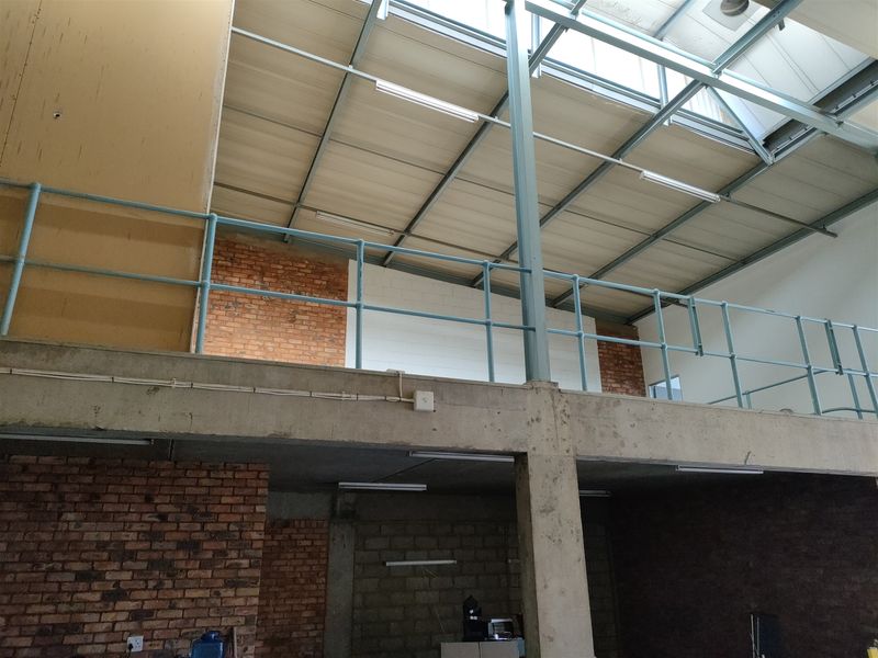 Industrial utility space featuring a mezzanine level with steel railings, exposed brick walls, concrete structural elements, and natural light provided by overhead skylights.