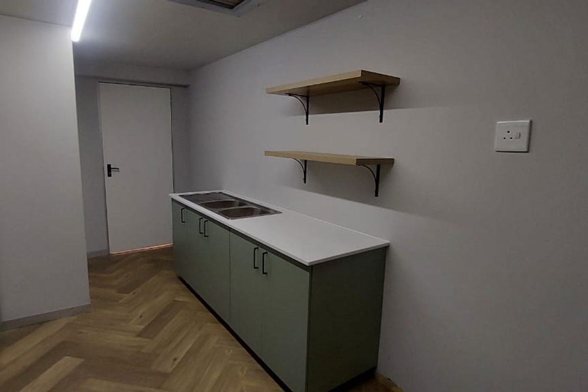 A clean, modern kitchen area featuring sage green cabinetry, a double sink, white countertops, and stylish herringbone wood-look flooring.