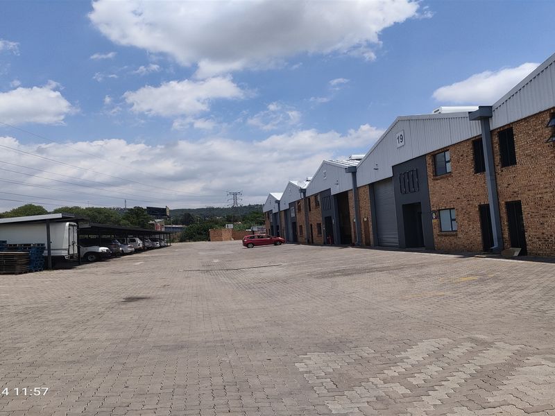 Exterior view of an industrial complex featuring multiple warehouse units with brick facades, metal roller shutter doors, and a large paved parking area.