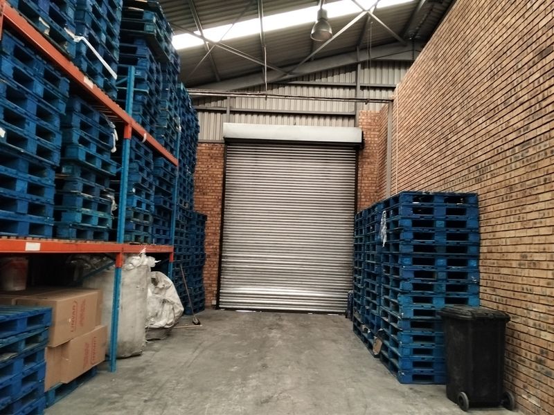 Industrial warehouse storage area featuring high ceilings with exposed steel trusses, face brick walls, concrete flooring, and a large roller shutter door.