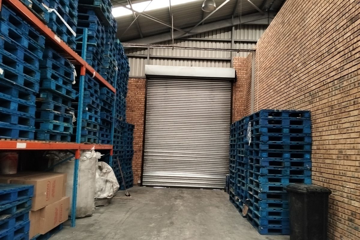 Industrial warehouse storage area featuring high ceilings with exposed steel trusses, face brick walls, concrete flooring, and a large roller shutter door.