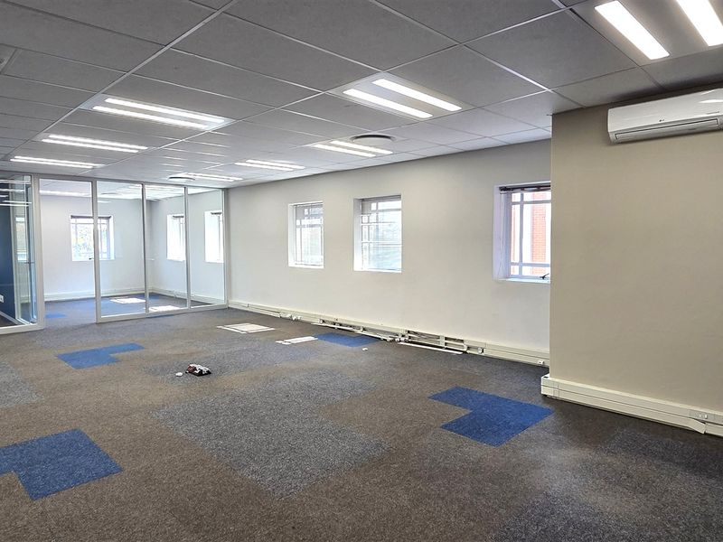 Spacious open-plan office area featuring modern glass partitions, suspended ceilings with recessed lighting, wall-mounted air conditioning, and durable carpet tile flooring.