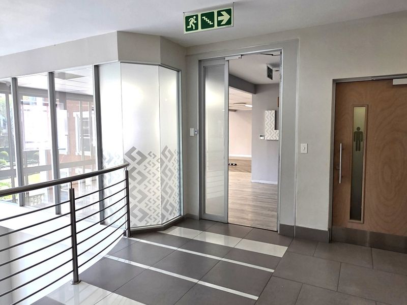A modern office hallway featuring tiled flooring, a stainless steel balustrade, and glass partitioning leading into an open-plan workspace.