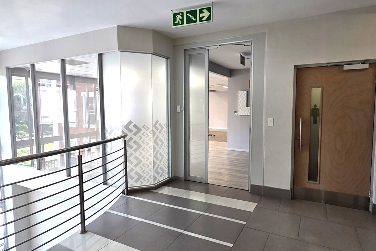 A modern office hallway featuring tiled flooring, a stainless steel balustrade, and glass partitioning leading into an open-plan workspace.