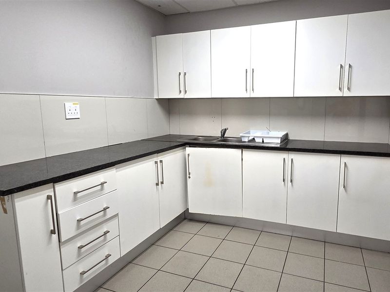 A functional office kitchenette featuring white laminate cabinetry, dark granite countertops, a stainless steel sink, and neutral ceramic floor tiling.