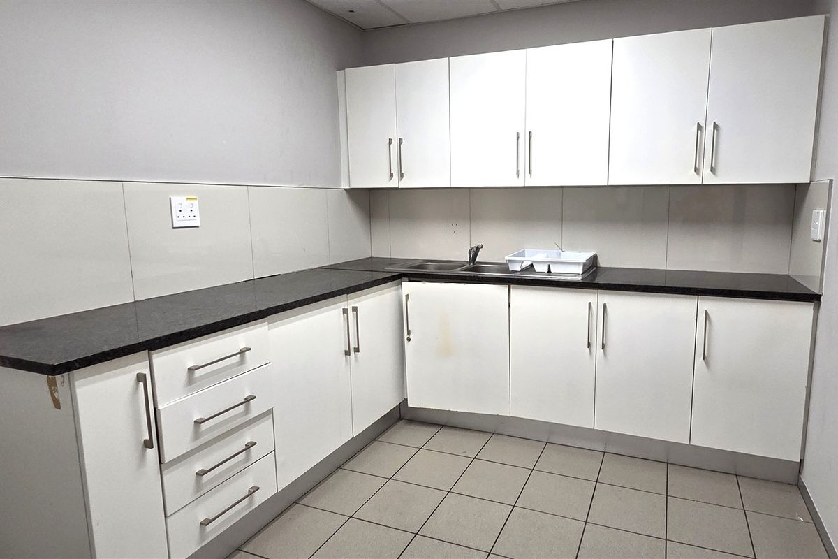 A functional office kitchenette featuring white laminate cabinetry, dark granite countertops, a stainless steel sink, and neutral ceramic floor tiling.