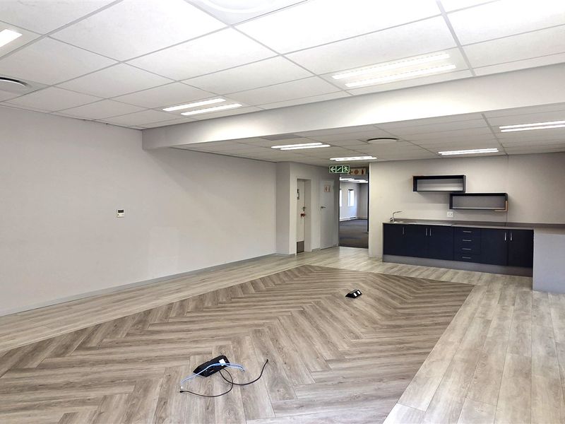 A spacious, modern open-plan office area featuring herringbone vinyl flooring, a functional kitchenette, and recessed ceiling lighting.