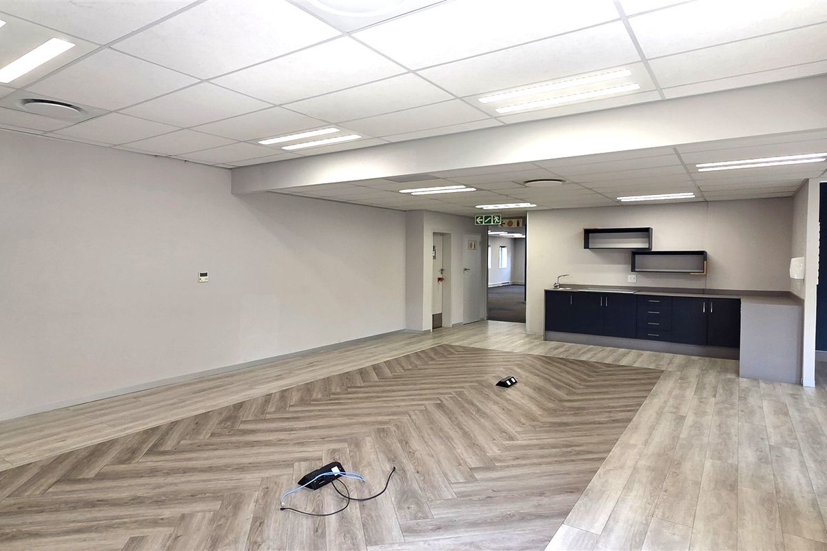 A spacious, modern open-plan office area featuring herringbone vinyl flooring, a functional kitchenette, and recessed ceiling lighting.