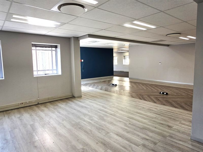 A spacious, open-plan office area featuring modern wood-look flooring, suspended ceilings with recessed lighting, and large windows with security bars.