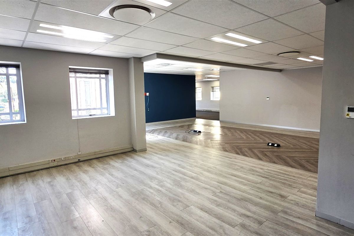 A spacious, open-plan office area featuring modern wood-look flooring, suspended ceilings with recessed lighting, and large windows with security bars.