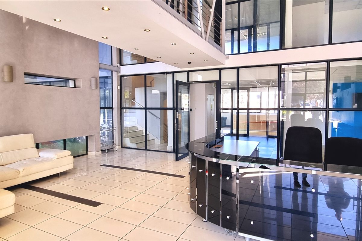 Modern office reception area featuring high ceilings, glass partitioning, tiled flooring, and a professional reception desk with a mezzanine level above.
