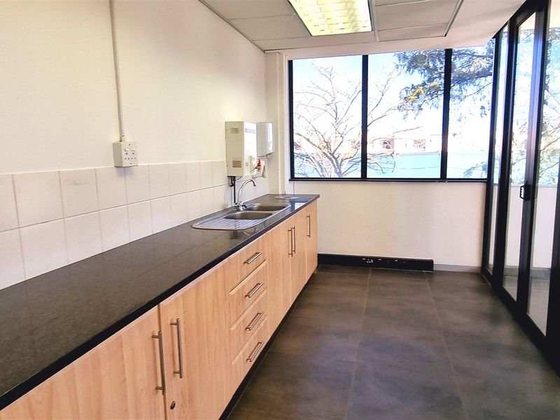 A clean, modern office kitchenette featuring light wood cabinetry, dark stone countertops, a stainless steel sink, and large windows providing natural light.