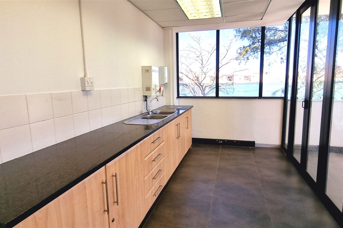 A clean, modern office kitchenette featuring light wood cabinetry, dark stone countertops, a stainless steel sink, and large windows providing natural light.