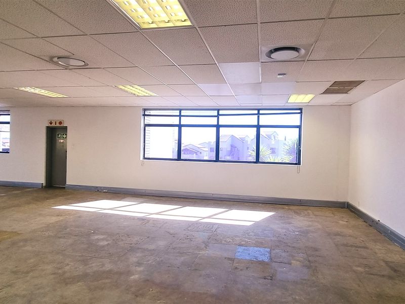 Spacious open-plan office area featuring a suspended ceiling, recessed lighting, and large windows providing ample natural light, with concrete flooring throughout.