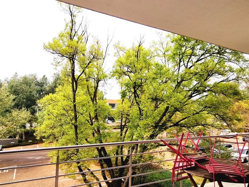 A spacious balcony featuring a stainless steel balustrade and tiled flooring, offering an elevated view of the surrounding trees and parking area.