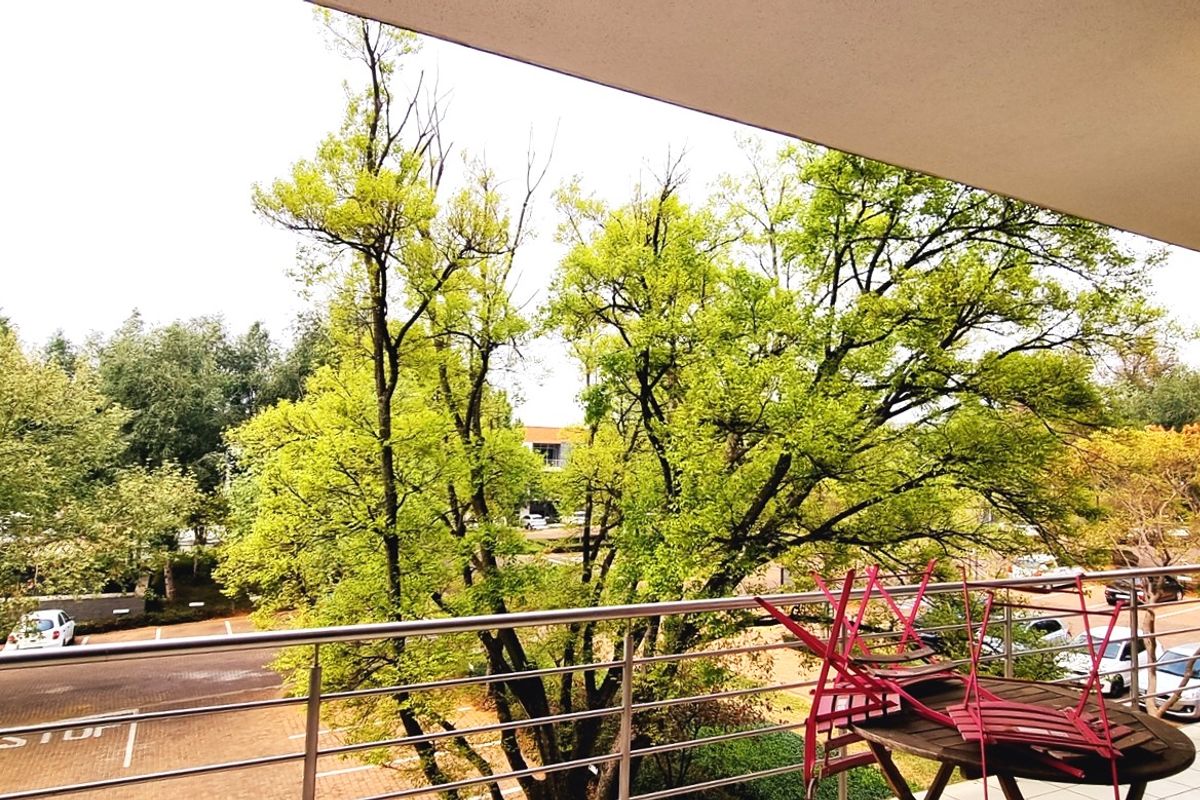 A spacious balcony featuring a stainless steel balustrade and tiled flooring, offering an elevated view of the surrounding trees and parking area.