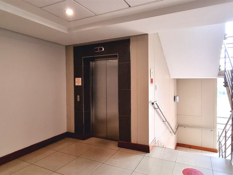 A clean and well-maintained office building hallway featuring a modern elevator, tiled flooring, and a staircase with metal handrails.