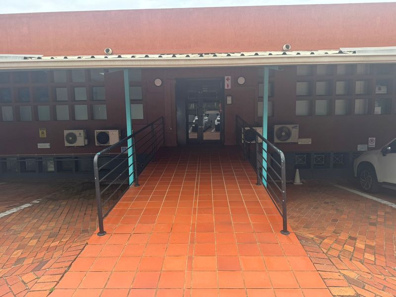 Office building entrance featuring a tiled accessibility ramp with metal handrails and wall-mounted air conditioning units.