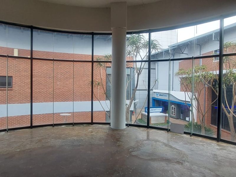An open-plan retail space featuring expansive floor-to-ceiling curved glass windows, a central structural column, and exposed concrete flooring overlooking an outdoor courtyard area.