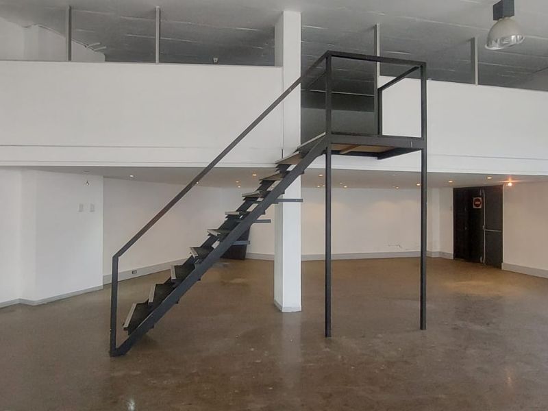 Open-plan retail space featuring a mezzanine level, industrial-style metal staircase, polished concrete flooring, and large glass storefront windows.