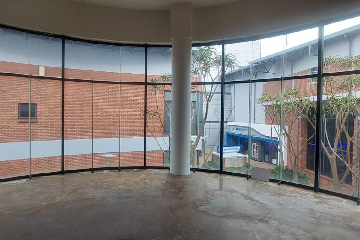 An open-plan retail space featuring expansive floor-to-ceiling curved glass windows, a central structural column, and exposed concrete flooring overlooking an outdoor courtyard area.