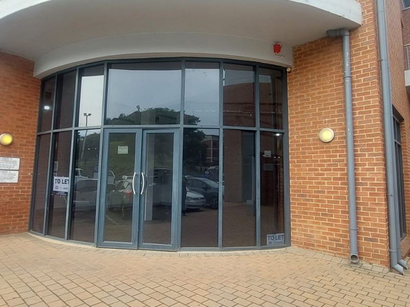 Front entrance of a commercial retail or office building featuring a curved glass facade, brick exterior, and paved walkway.
