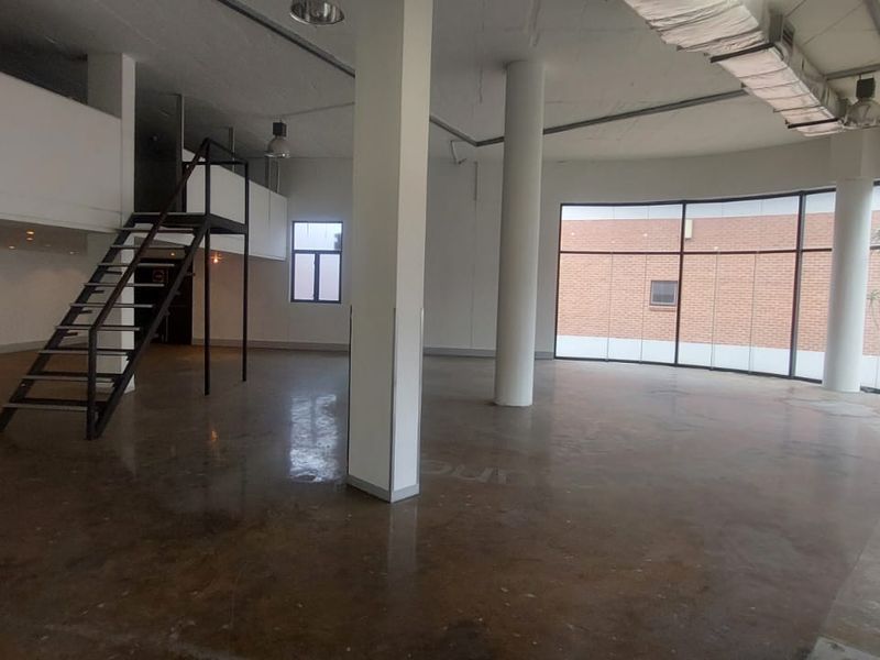 Spacious open-plan retail or office area featuring polished concrete floors, a mezzanine level with metal staircase, and large curved floor-to-ceiling windows.