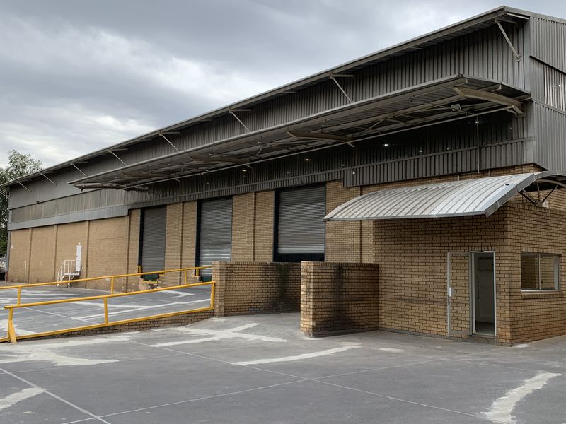 Industrial warehouse exterior featuring a concrete loading yard, multiple roller shutter doors, brick facade, and metal cladding with a protective awning.