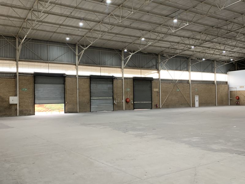 Spacious industrial warehouse featuring high ceilings, exposed steel trusses, concrete flooring, and multiple roller shutter doors for efficient loading and access.