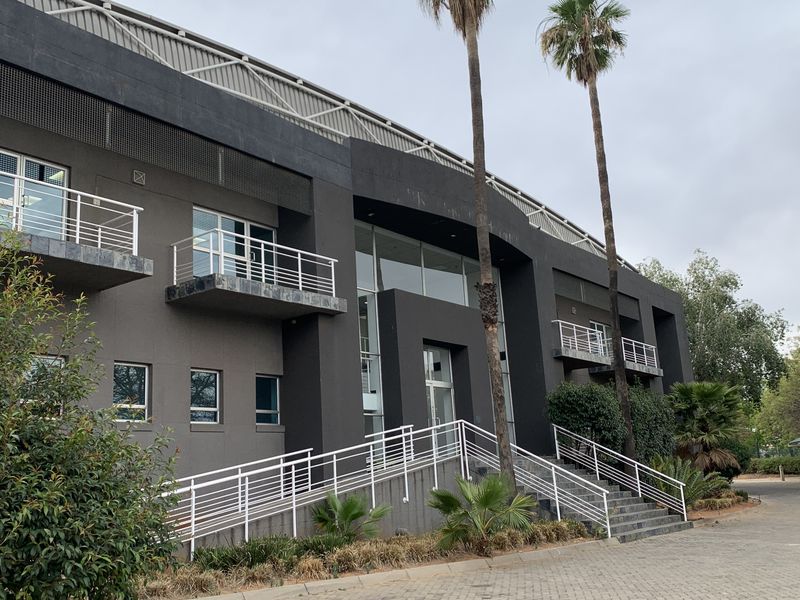 Modern industrial office building facade featuring a prominent entrance, wheelchair ramp, concrete staircase, and multiple balconies with metal railings.