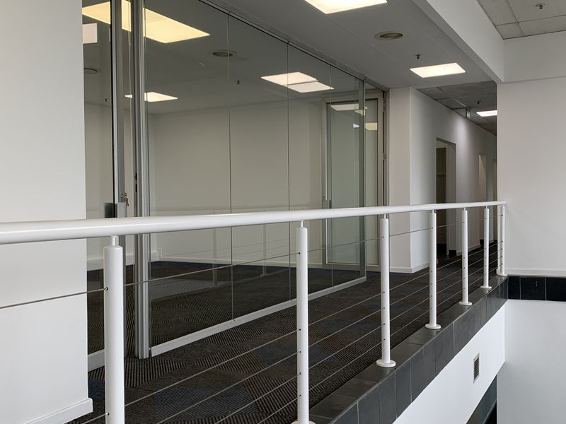 A bright, modern industrial hallway featuring glass partition walls, recessed lighting, and a clean metal railing overlooking the lower level.