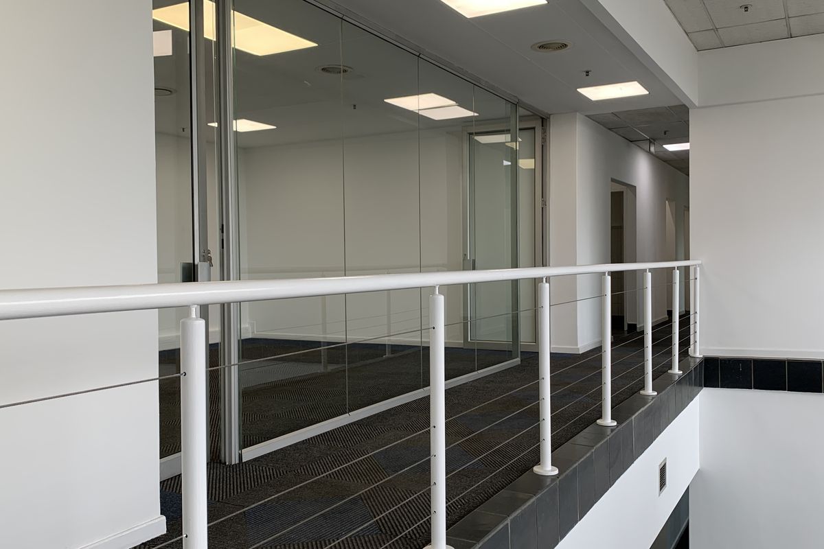 A bright, modern industrial hallway featuring glass partition walls, recessed lighting, and a clean metal railing overlooking the lower level.