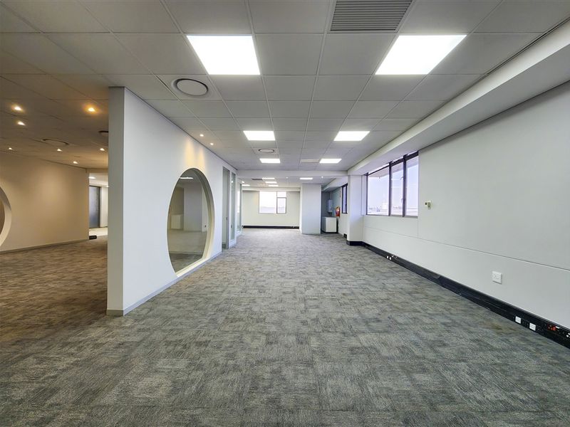 A spacious, open-plan office area featuring modern suspended ceilings, recessed LED lighting, carpet tiles, and a unique circular interior window design.