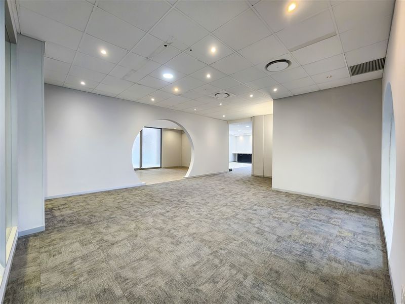 A spacious, vacant office area featuring a modern circular archway, neutral carpet tiles, and a suspended ceiling with recessed lighting.