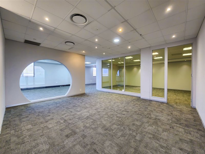 Modern open-plan office space featuring carpet tiles, suspended ceilings with recessed lighting, and glass partitions with a unique circular window design.
