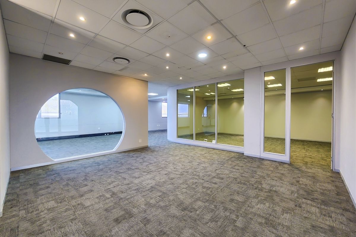 Modern open-plan office space featuring carpet tiles, suspended ceilings with recessed lighting, and glass partitions with a unique circular window design.