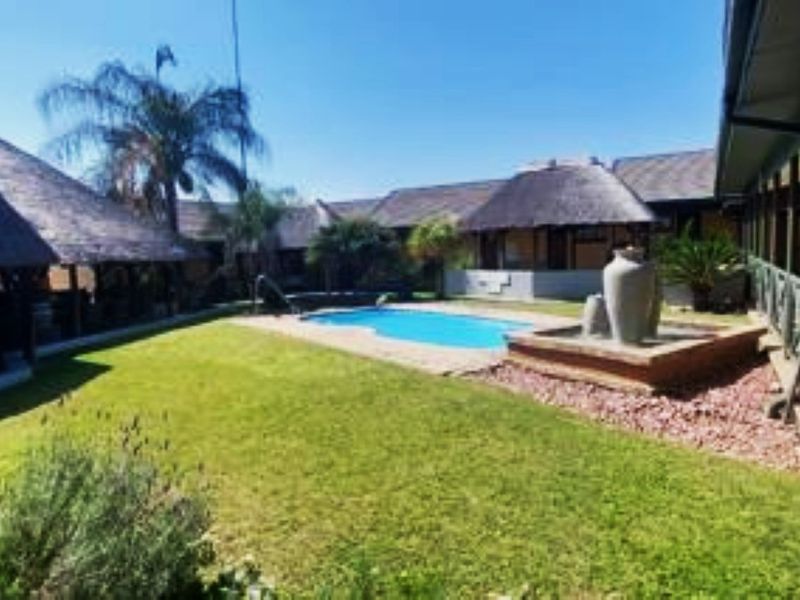 A well-maintained lodge courtyard featuring a swimming pool, central water feature, and surrounding thatched-roof structures set within a landscaped garden.